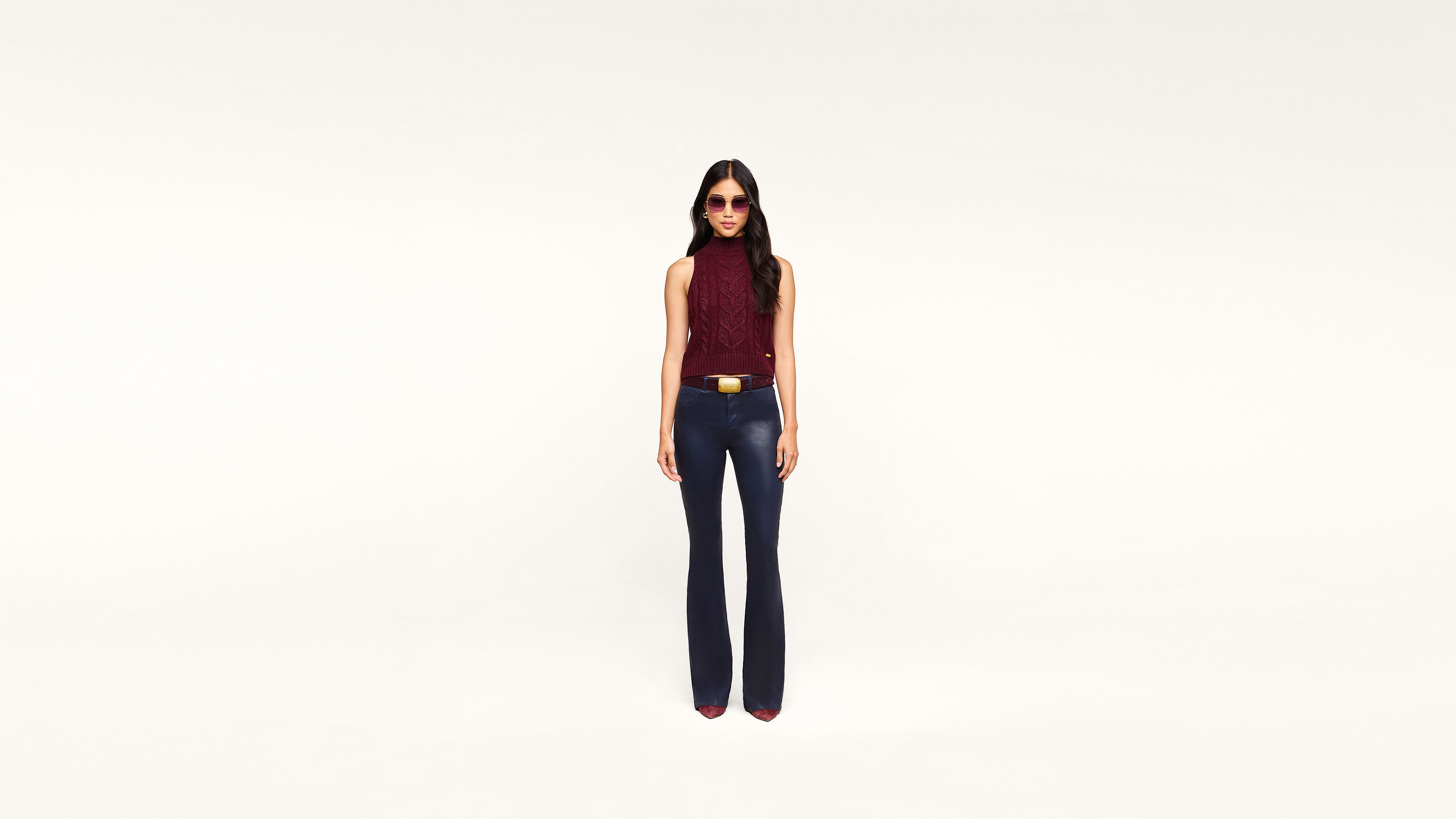 A woman wearing sunglasses, a sleeveless maroon top, dark pants, and red shoes stands confidently against a plain white background.