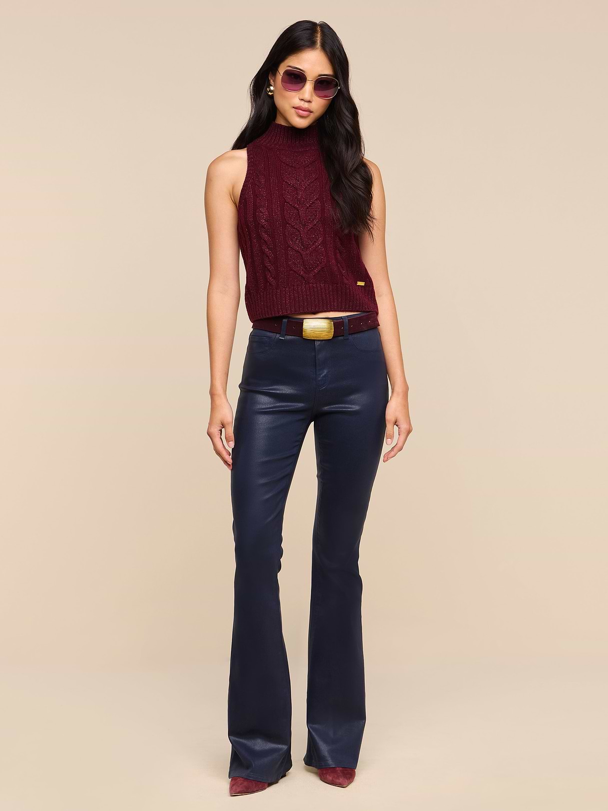 A woman wearing sunglasses, a sleeveless burgundy turtleneck sweater, a brown belt with a large buckle, pointed burgundy shoes, and the Marty Coated Flare Jean stands against a plain white background.