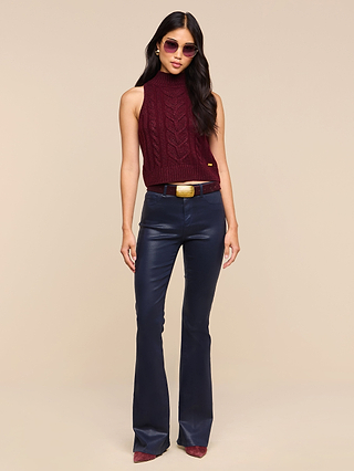Woman wearing sunglasses, a sleeveless maroon knit top, a black belt with a gold buckle, the Marty Coated Flare Jean in a high rise dark finish, and maroon shoes, standing against a beige background.