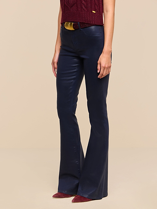 A person wearing a sleeveless burgundy top, the Marty Coated Flare Jean in high-rise navy blue with a flared hem, a brown belt with a gold buckle, and burgundy pointed shoes stands against a plain beige background.