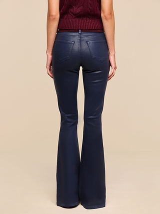 A person stands facing away, wearing a sleeveless burgundy knit top and the Marty Coated Flare Jean in dark blue high rise with a subtle flared hem. The background is plain beige.