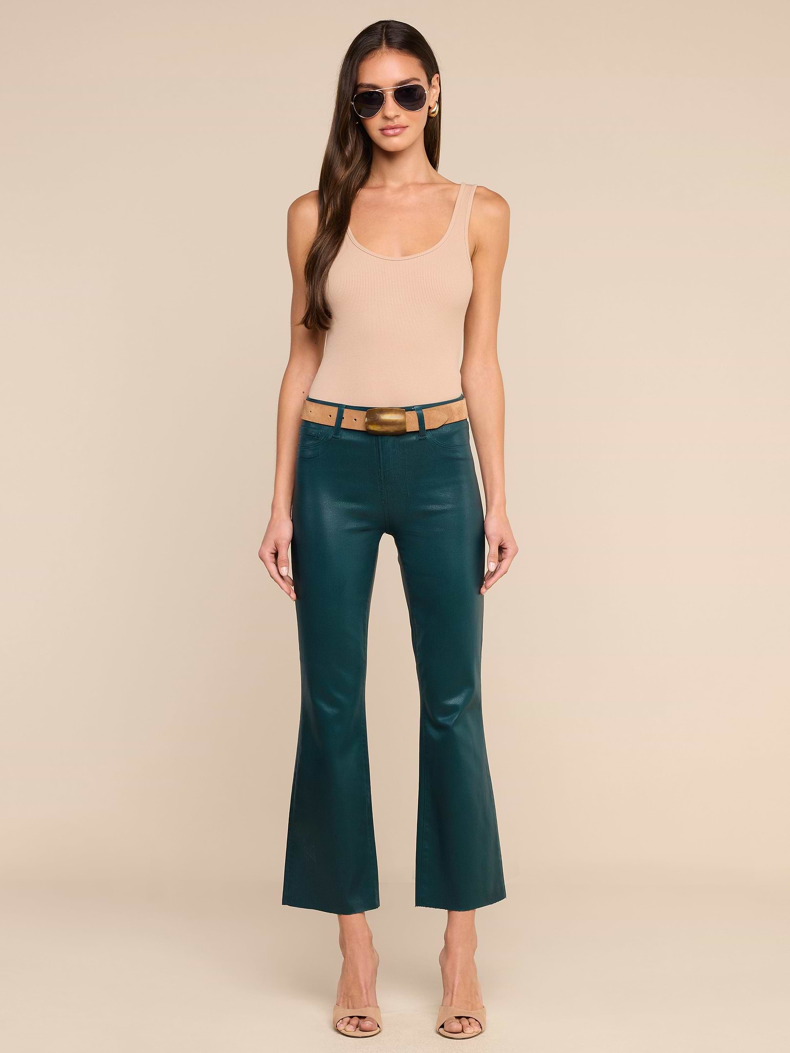 A woman stands against a plain white background wearing sunglasses, a beige sleeveless top, the Kendra Coated Cropped Flare Jean in dark green with a tan belt, and beige heels.