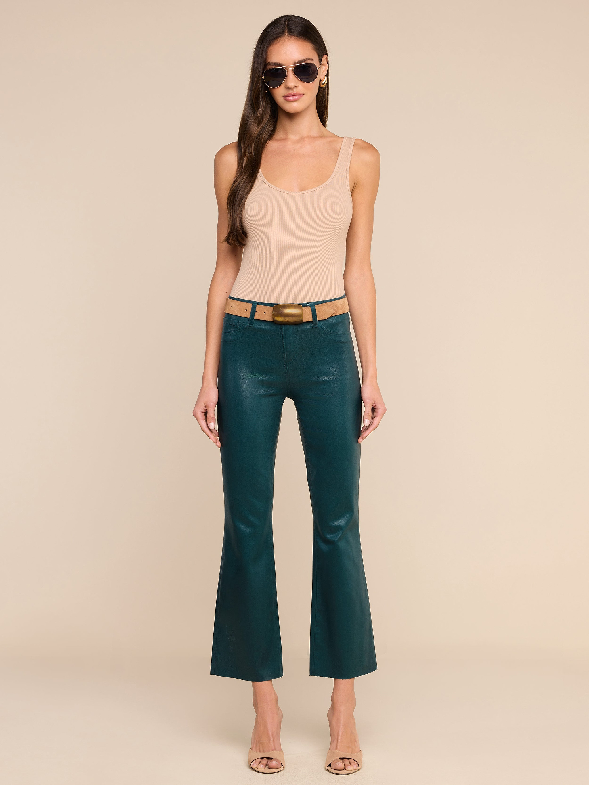 Une femme se tient debout devant un fond beige ; elle porte des lunettes de soleil, un haut sans manches beige, des sandales à talons beiges et le « Kendra Coated Cropped Flare Jean » — un jean évasé court à taille haute de couleur bleu sarcelle, muni dune ceinture.