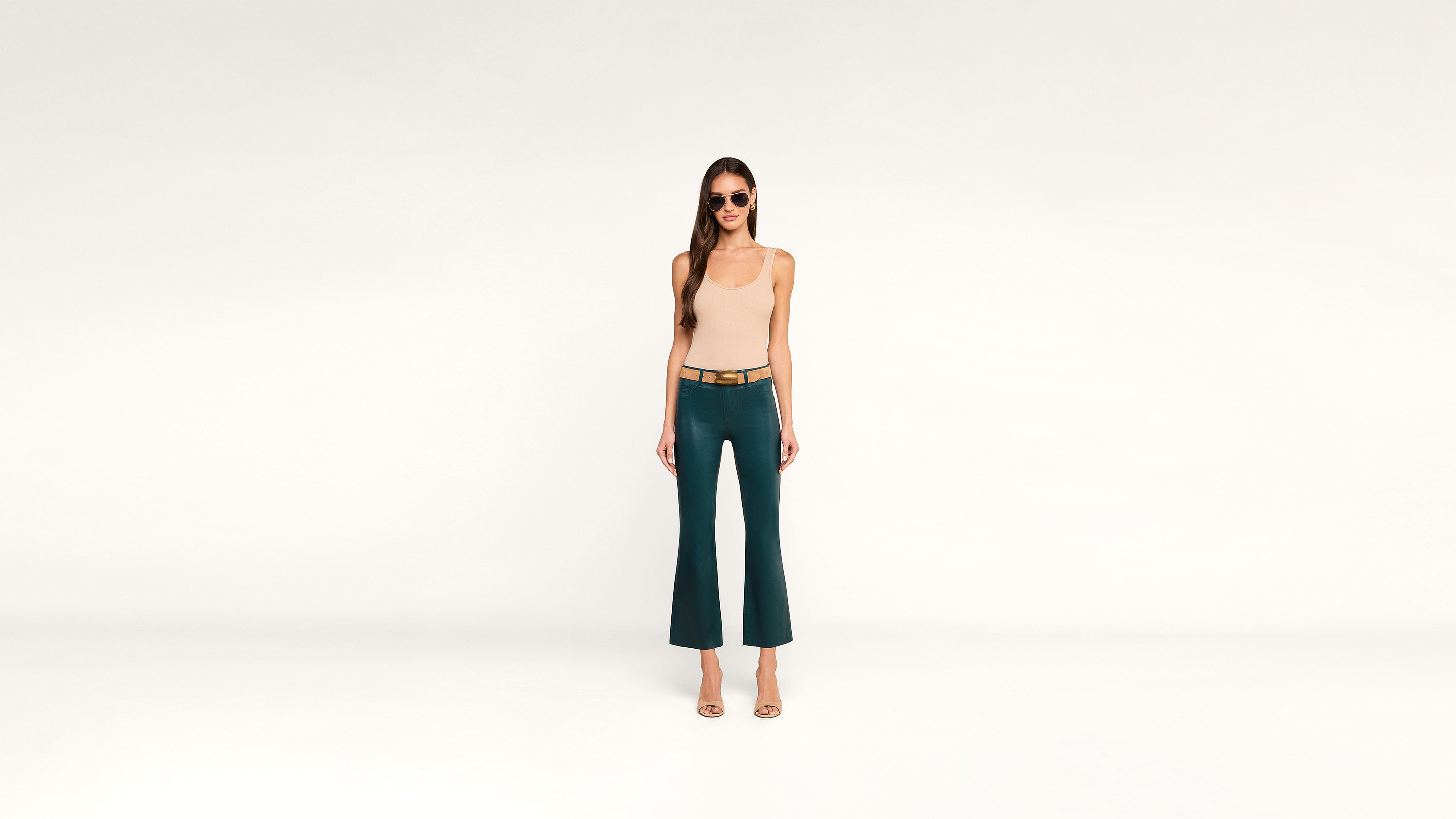 A woman wearing sunglasses, a beige sleeveless top, and teal pants stands on a white background, facing forward with her arms relaxed by her sides.