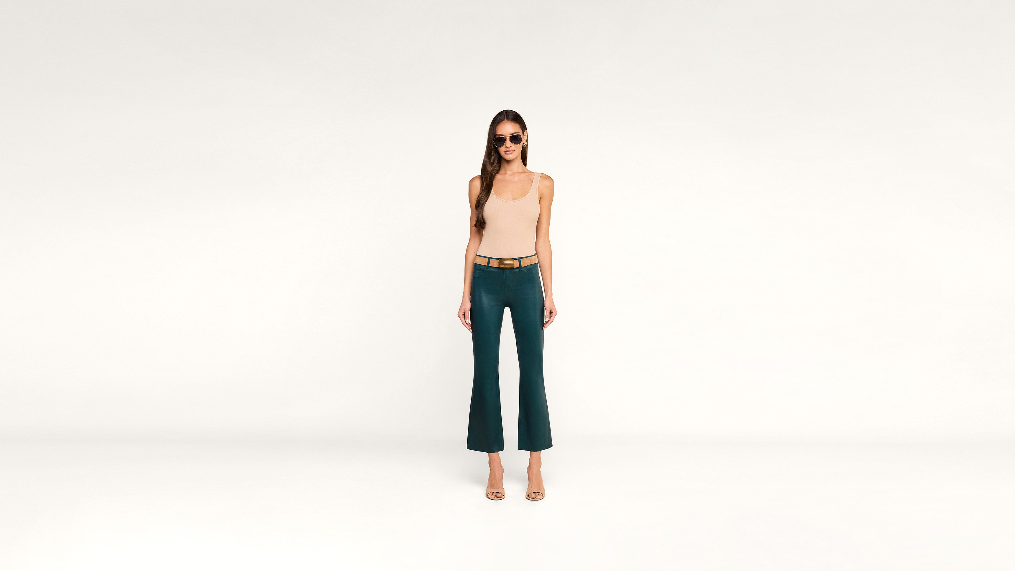 A woman wearing sunglasses, a beige sleeveless top, and teal pants stands on a white background, facing forward with her arms relaxed by her sides.