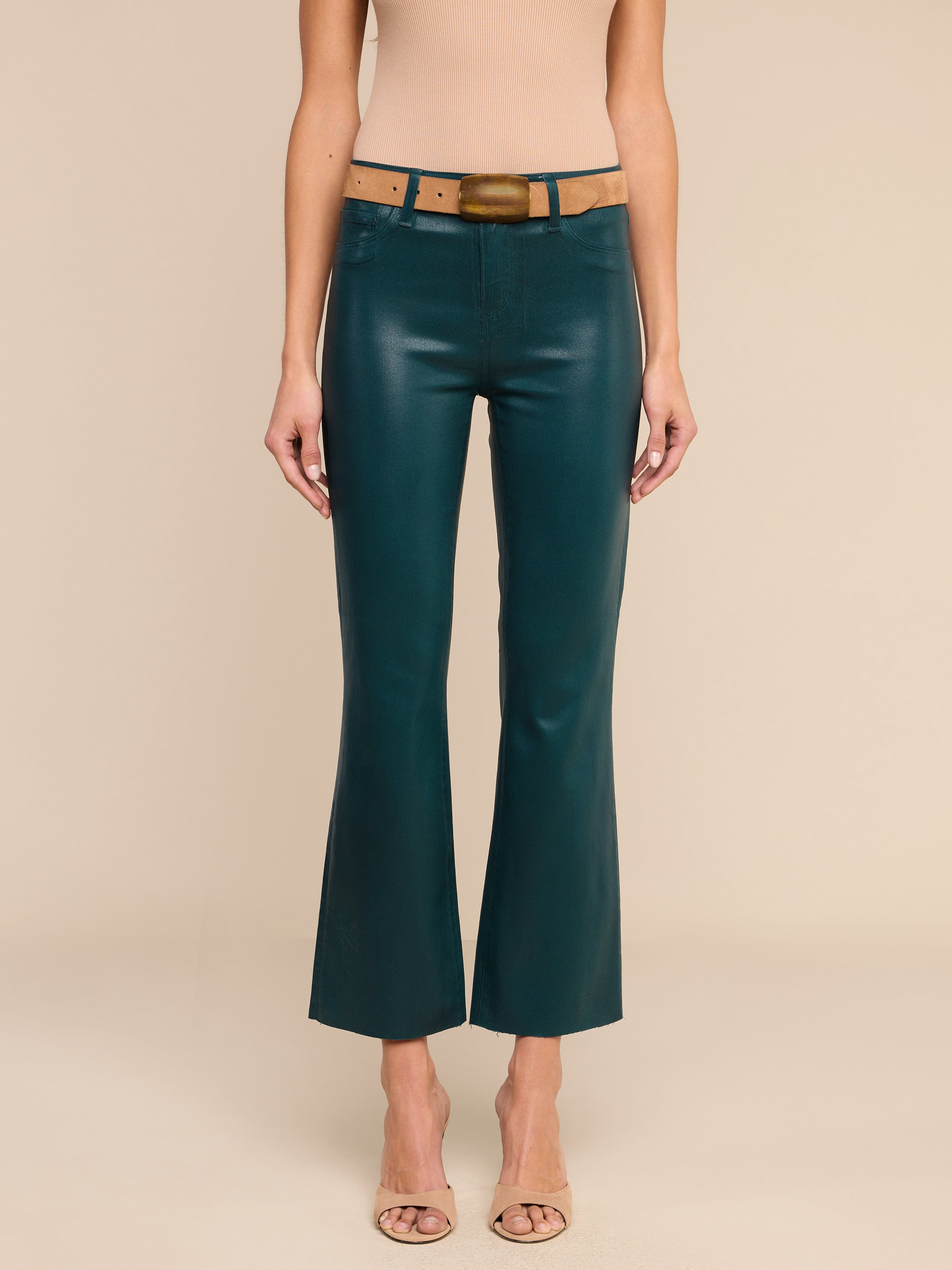 Une personne portant un jean évasé court enduit « Kendra » de couleur bleu sarcelle, une ceinture beige clair, un haut beige sans manches et des escarpins nude à bout ouvert, se tenant debout devant un fond beige uni.