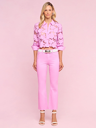 Una donna dai capelli biondi e dallespressione neutra è in piedi su uno sfondo rosa e indossa un paio di jeans Kendra Coated Cropped Flare, una camicetta rosa chiaro con pizzo a forma di farfalla, una cintura bianca e sandali bianchi con tacco.