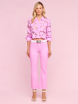 A woman with blonde hair and a neutral expression stands against a pink background wearing Kendra Coated Cropped Flare Jeans, a light pink lacy butterfly blouse, a white belt, and white heeled sandals.
