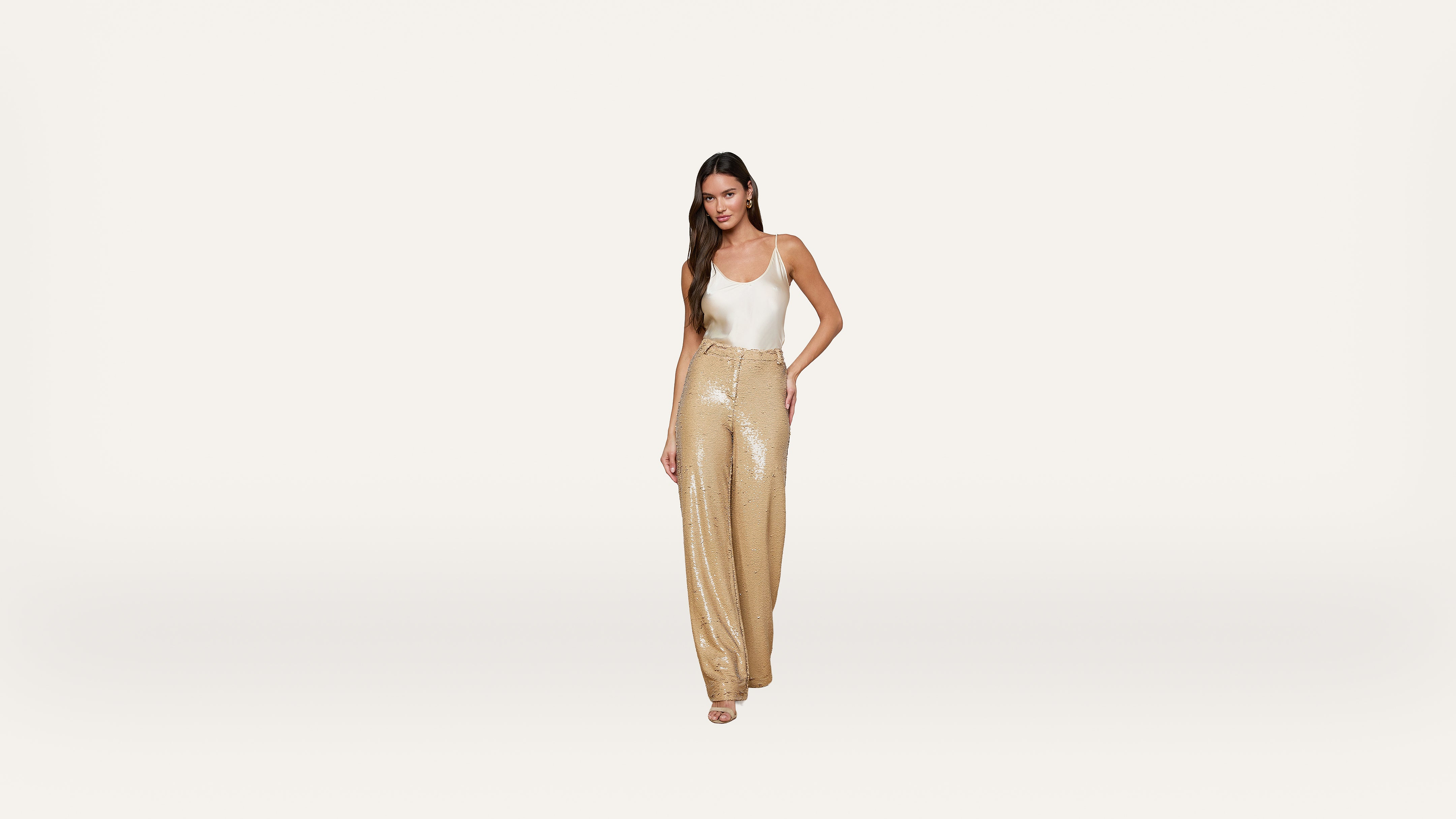 Woman in a white tank top and gold sequin wide-leg pants poses confidently against a plain white background.