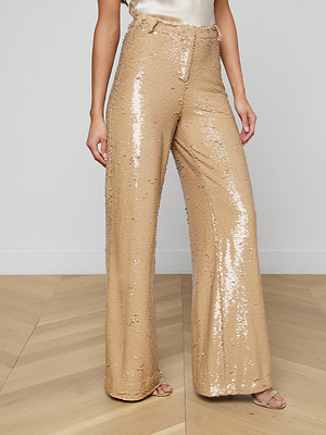 Someone stands on a light wood floor against a plain white wall, wearing the Gavin Sequin Wide-Leg Pant in gold with open-toe heels. Their high-rise pants shimmer, and their top is partially visible.