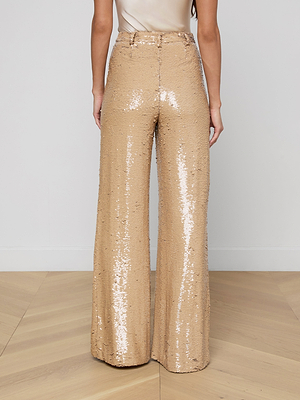 A person faces away in the Gavin Sequin Wide-Leg Pant, featuring high-rise, wide-leg gold sequins that shimmer and reflect light onto a wooden floor against a plain white background.