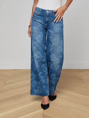 A person stands on a light wood floor wearing Alicent Wide-Leg Jeans with a floral print, paired with a white top and black pointed-toe shoes. The image is cropped at the shoulders.