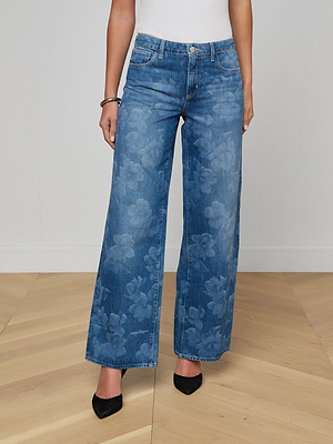 A person wears the Alicent Wide-Leg Jean in blue with a subtle floral pattern, paired with a white top, black pointed-toe shoes, and a bracelet, standing on a wooden floor against a plain white wall.