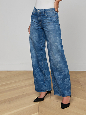 A person wearing Alicent Wide-Leg Jean in a subtle floral print and black high-heeled shoes stands on a light wood floor against a plain white wall.