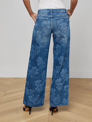 A person stands on a wooden floor, wearing the Alicent Wide-Leg Jean at sneaker length with black high-heeled sandals. The photo, taken from behind, highlights the back pockets and detailed floral print.