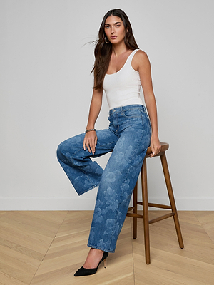 A woman with long brown hair sits on a wooden stool, wearing a white sleeveless top, black high heels, and the Alicent Wide-Leg Jean in a floral print. The background is a plain white wall with a light wood floor.