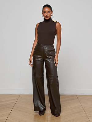 A woman stands indoors against a plain wall, wearing a sleeveless brown turtleneck top with gold buttons and high-waisted Livvy Leather Trousers by L'AGENCE. Her hair is pulled back and she pairs the look with heels.