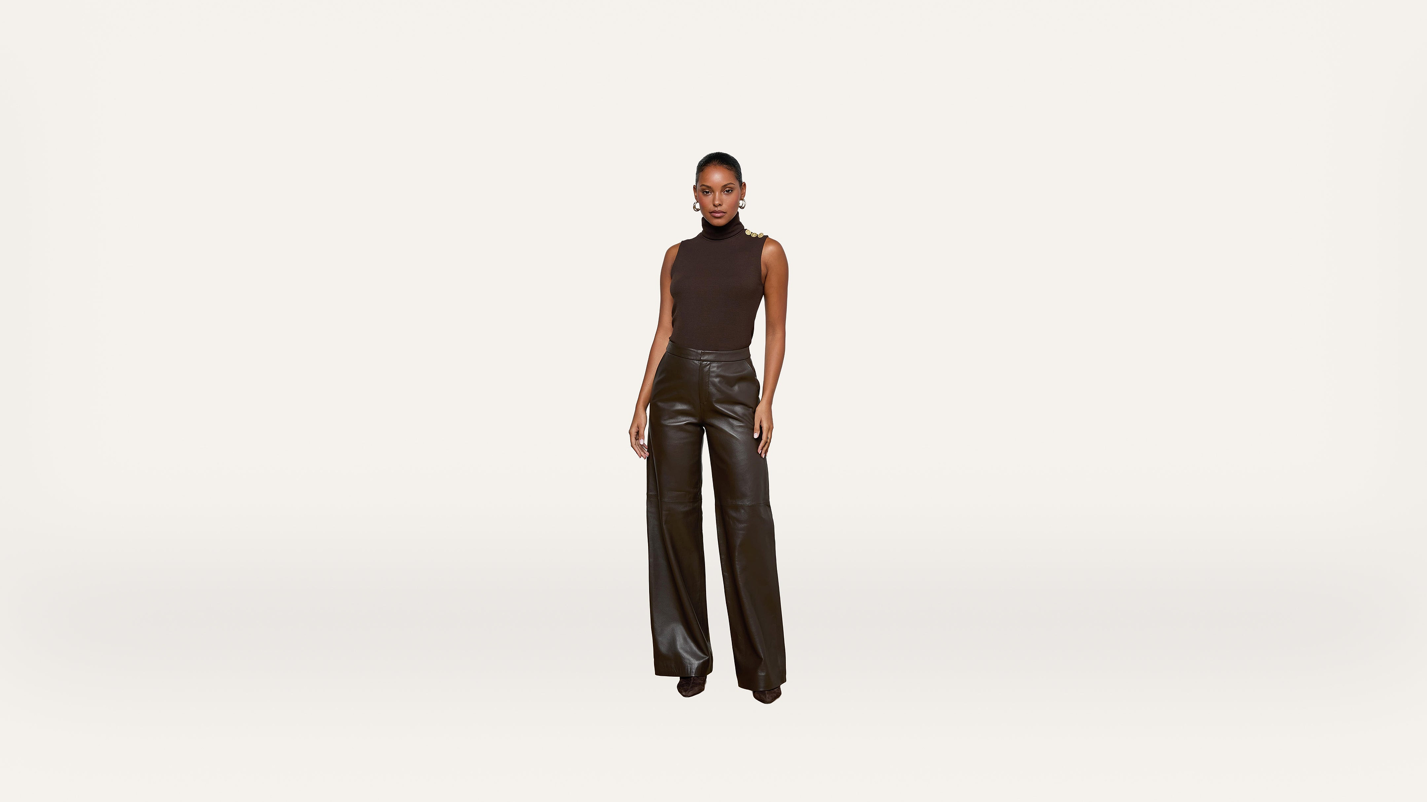A woman stands facing forward, wearing a sleeveless black top and high-waisted, wide-leg black leather pants against a plain white background.