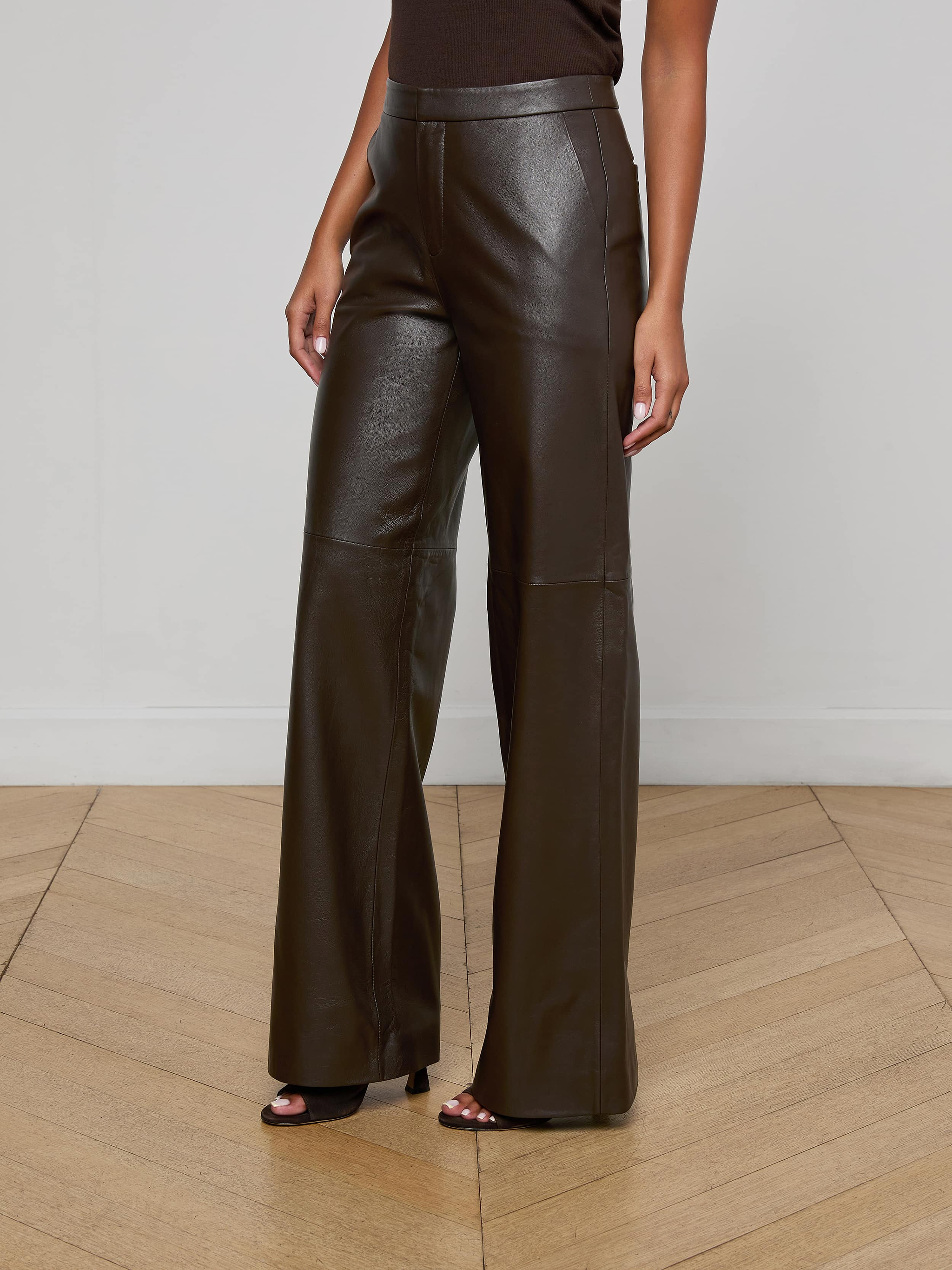 A person wears the L'AGENCE Livvy Leather Trouser—high-waisted, wide-leg chocolate pants—with a sleeveless black top, standing on a light wood herringbone floor. Their arms are relaxed by their sides, and their face is not visible.