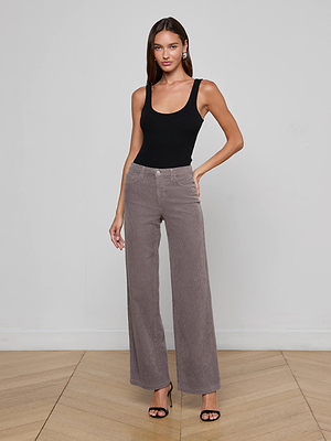 A woman stands indoors against a plain light wall, wearing a black tank top and high-waisted taupe Scottie Corduroy Wide-Leg Jeans with a contoured waistband. She completes the look with black heeled sandals and her long brown hair frames a slight smile.