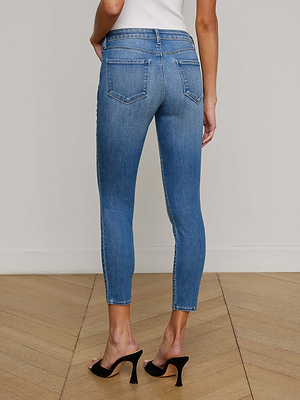 A person wearing L'AGENCE Frankie Low-Rise Skinny Jean in light blue and black high-heeled sandals stands on a wooden floor, facing away from the camera against a plain white wall.