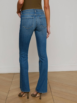 A person wearing L'AGENCE Cara Low-Rise Bootcut Jeans and heeled sandals stands on a wooden floor, facing away from the camera with hands by their sides.