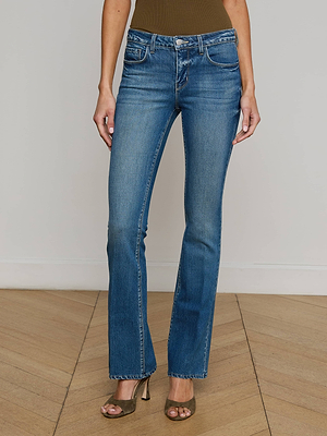 A person wearing L'AGENCE Cara Low-Rise Bootcut Jeans and olive green heeled sandals stands on a wooden floor, shown from the shoulders down.
