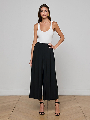 A woman poses confidently indoors against a light wall, wearing a white sleeveless top and L'AGENCE Novalynn Cropped Pleated Pant in black with black heeled sandals. She has long hair and radiates relaxed elegance.