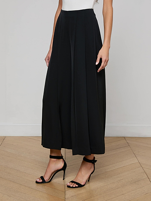 A person wearing a white sleeveless top, black high-heeled sandals, and the L'AGENCE Novalynn Cropped Pleated Pant stands on a wooden floor against a plain light-colored wall.