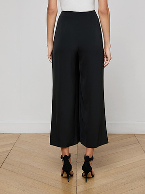 A woman stands indoors on a wooden floor, facing away from the camera, wearing L'AGENCE's Novalynn Cropped Pleated Pant in black, paired with a white sleeveless top and black open-toe heels.