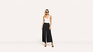A woman in sunglasses, a white sleeveless top, black wide-leg pants, and black heels stands confidently against a plain white background.