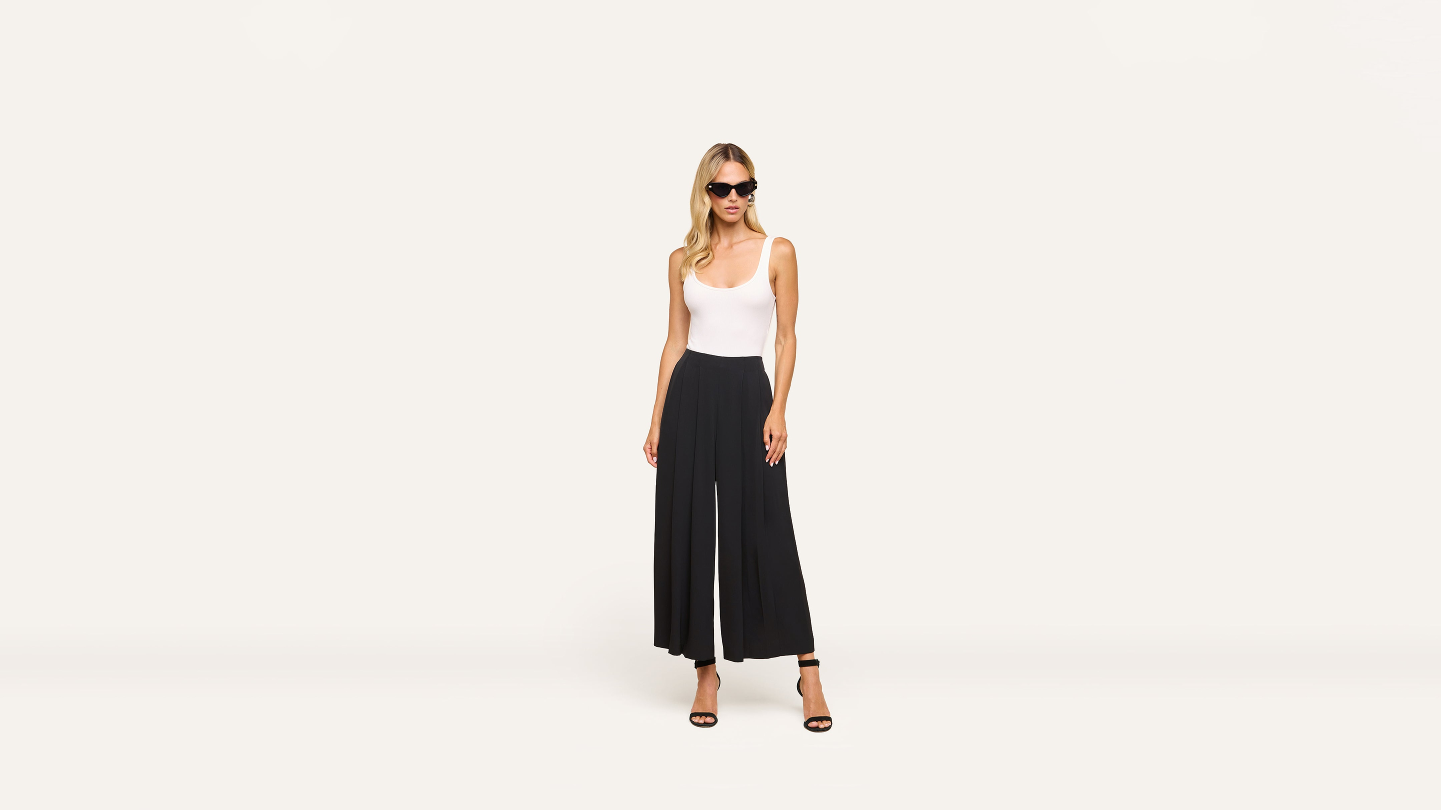 A woman in sunglasses, a white sleeveless top, black wide-leg pants, and black heels stands confidently against a plain white background.