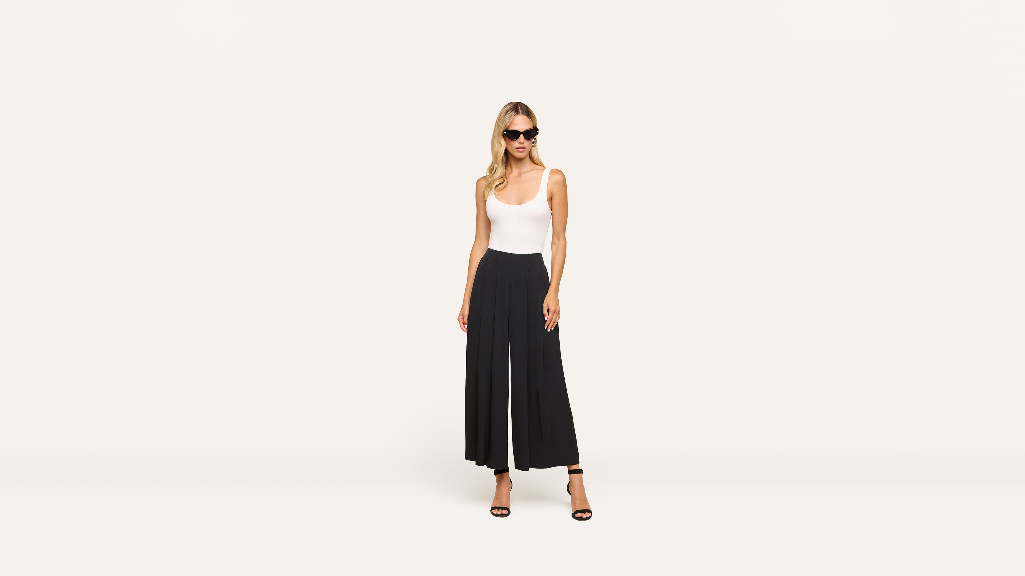 A woman in sunglasses, a white sleeveless top, black wide-leg pants, and black heels stands confidently against a plain white background.