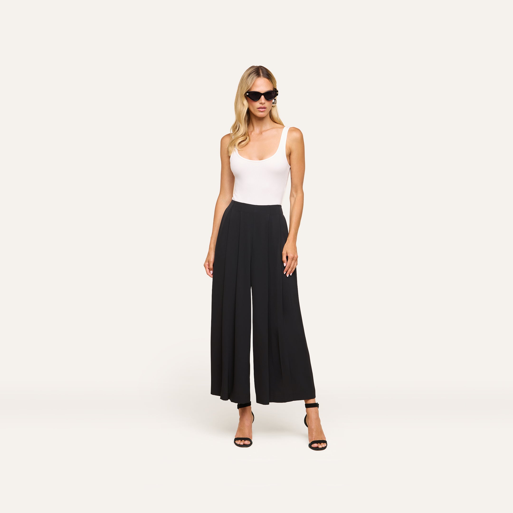 Woman wearing sunglasses, a white sleeveless top, Novalynn Cropped Pleated Pant in black, and black heeled sandals stands against a plain light background.