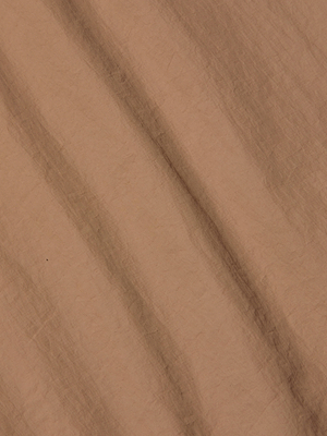 A close-up of light brown, crumpled paper with diagonal creases and textured lines creates a subtle striped pattern, echoing the lightweight nylon look of the Mariko Jogger.