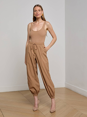 A woman indoors wears the beige Mariko Jogger, featuring a drawstring waist and lightweight nylon. Paired with nude heels and her hair down, she poses against a light wall and wooden floor.