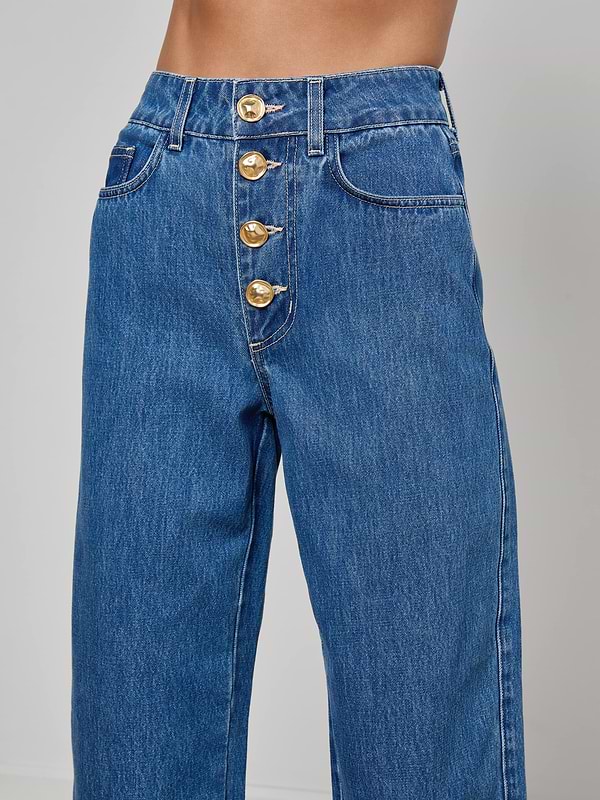 Close-up of a person wearing the Liora Cropped Jean, vintage-inspired high rise blue jeans featuring four large gold buttons and side and front pockets, standing against a plain light gray background.