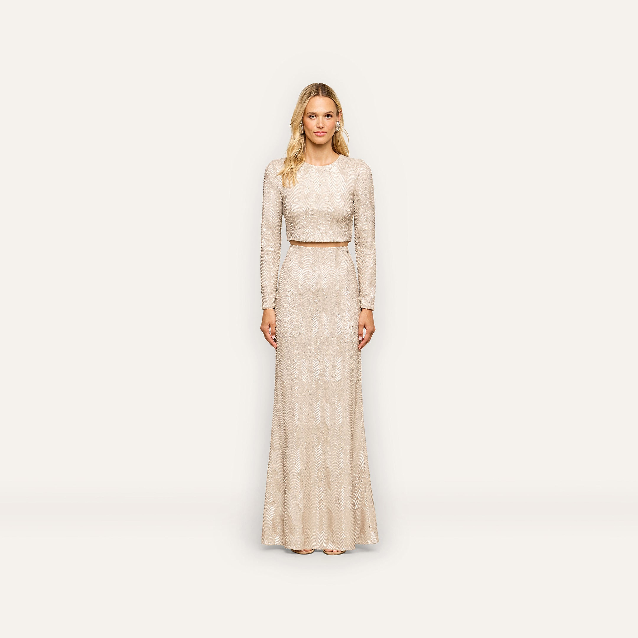 A woman stands against a plain white background, wearing the Zeta Sequin Maxi Skirt—a long, light-colored skirt adorned with subtle champagne-inspired sequin patterns. With her blonde hair and neutral expression, she presents an elegant look perfect for any special occasion.