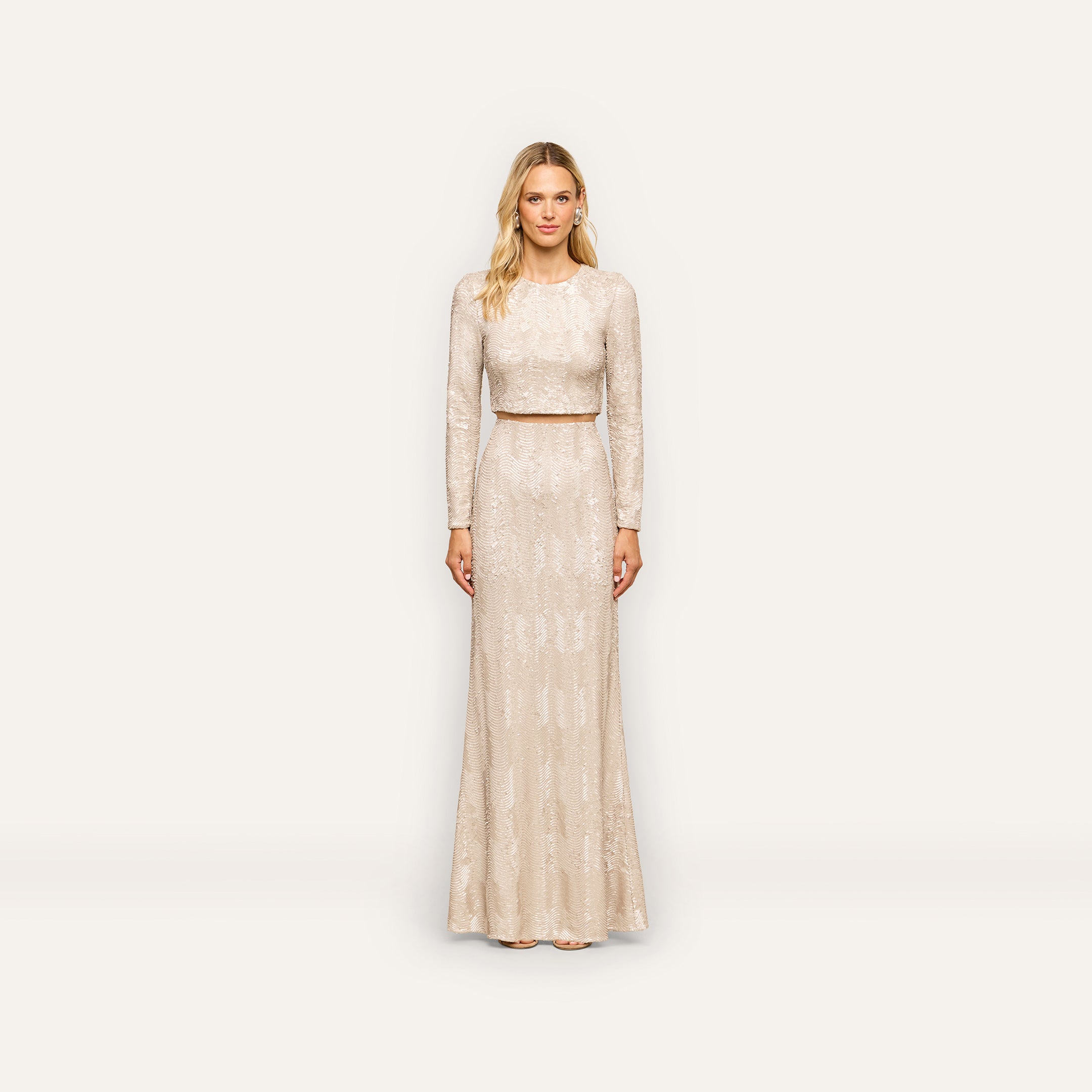 A woman stands against a plain white background, wearing the Zeta Sequin Maxi Skirt—a long, light-colored skirt adorned with subtle champagne-inspired sequin patterns. With her blonde hair and neutral expression, she presents an elegant look perfect for any special occasion.
