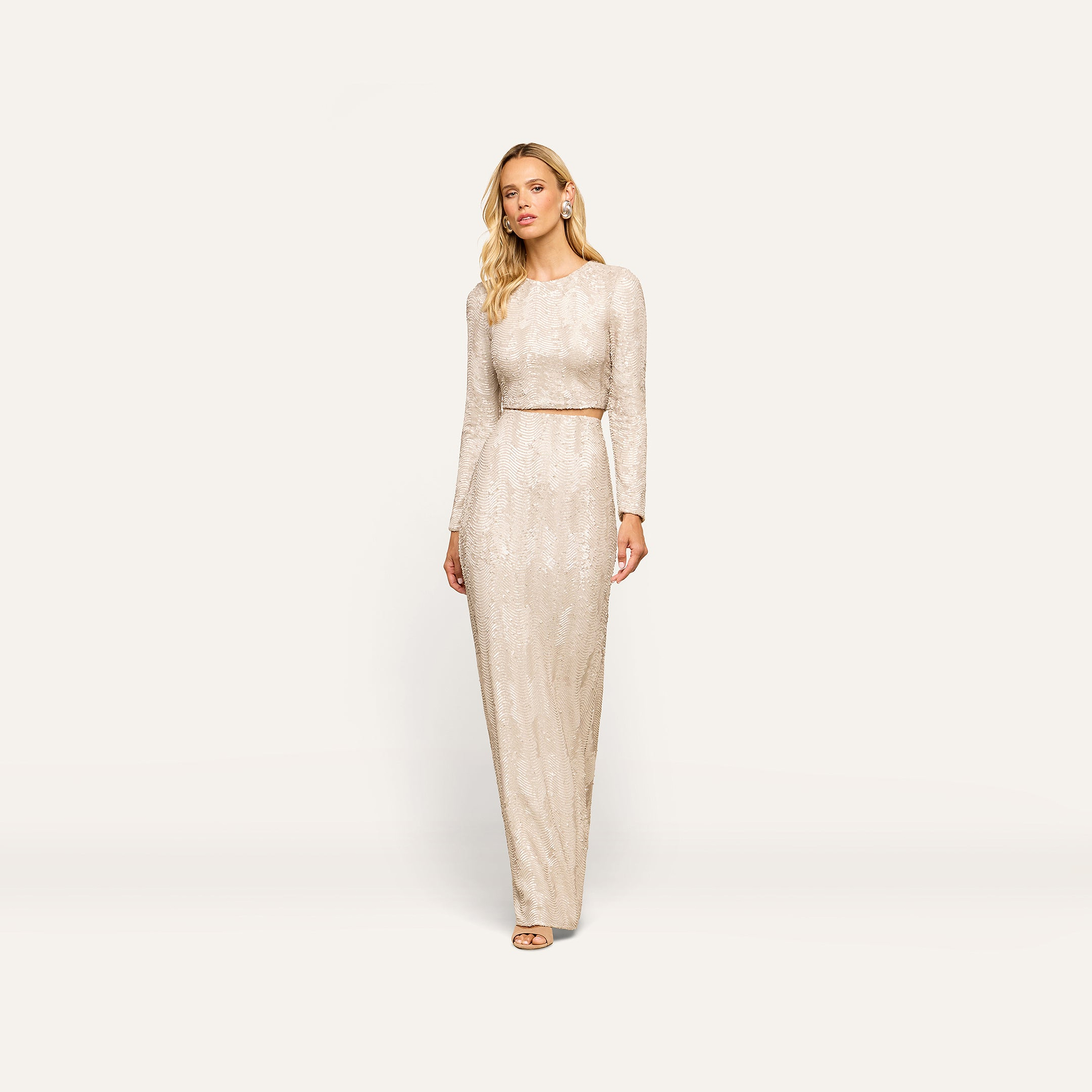 A woman models the Zeta Sequin Maxi Skirt, a floor-length design covered in shimmering champagne sequins, paired here with a coordinating long-sleeved top. She stands against a plain white background, gazing thoughtfully to the side. Perfect for special occasions, this skirt showcases elegant detailing inspired by the signature Zeta style.