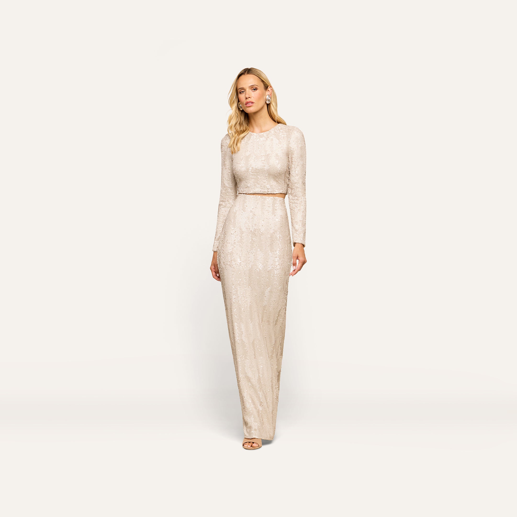 A woman models the Zeta Sequin Maxi Skirt, a floor-length design covered in shimmering champagne sequins, paired here with a coordinating long-sleeved top. She stands against a plain white background, gazing thoughtfully to the side. Perfect for special occasions, this skirt showcases elegant detailing inspired by the signature Zeta style.