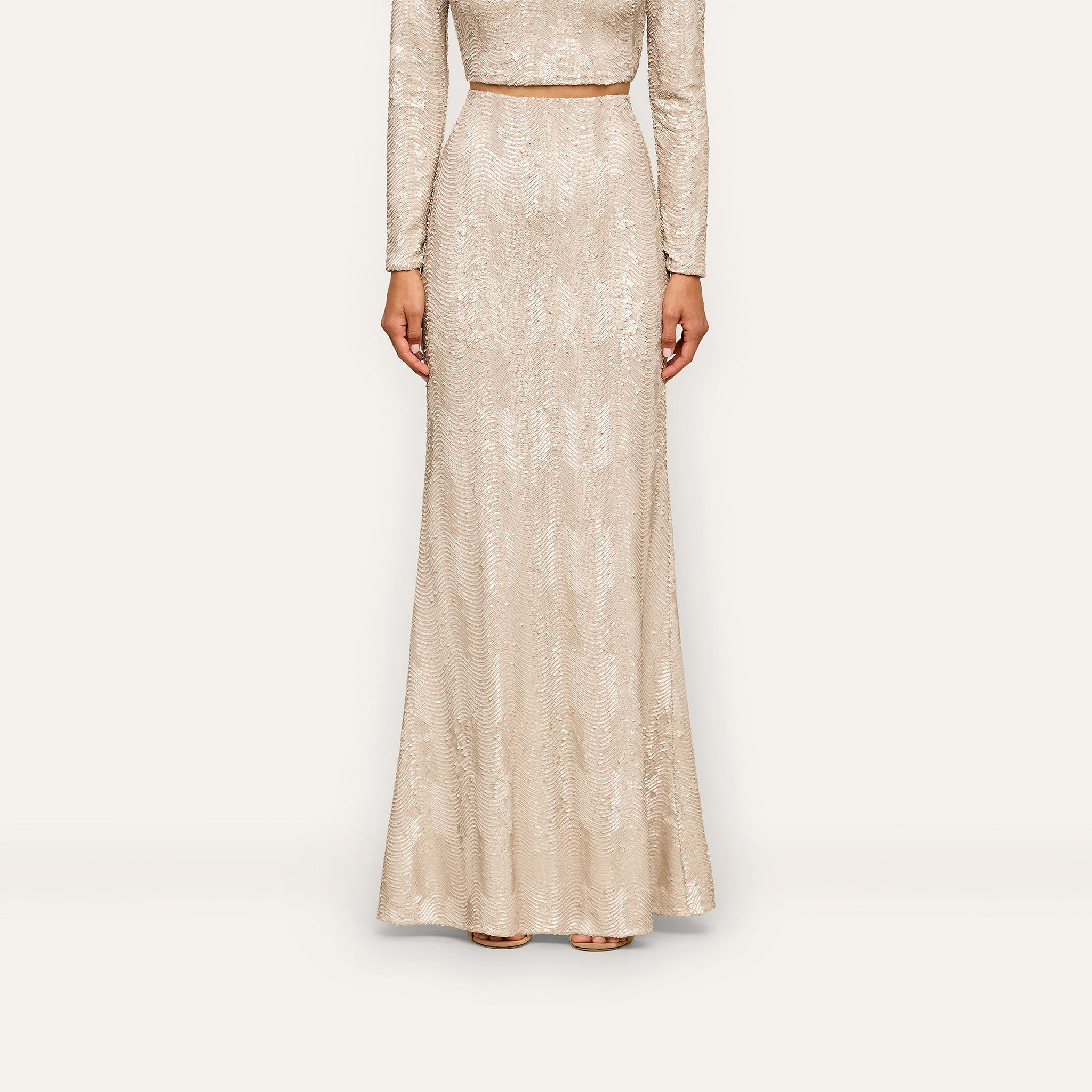 A person wearing the Zeta Sequin Maxi Skirt—a long, fitted, light beige skirt adorned with champagne sequins—stands against a plain white background. Only the body from the shoulders down is visible, making it a perfect look for a special occasion.