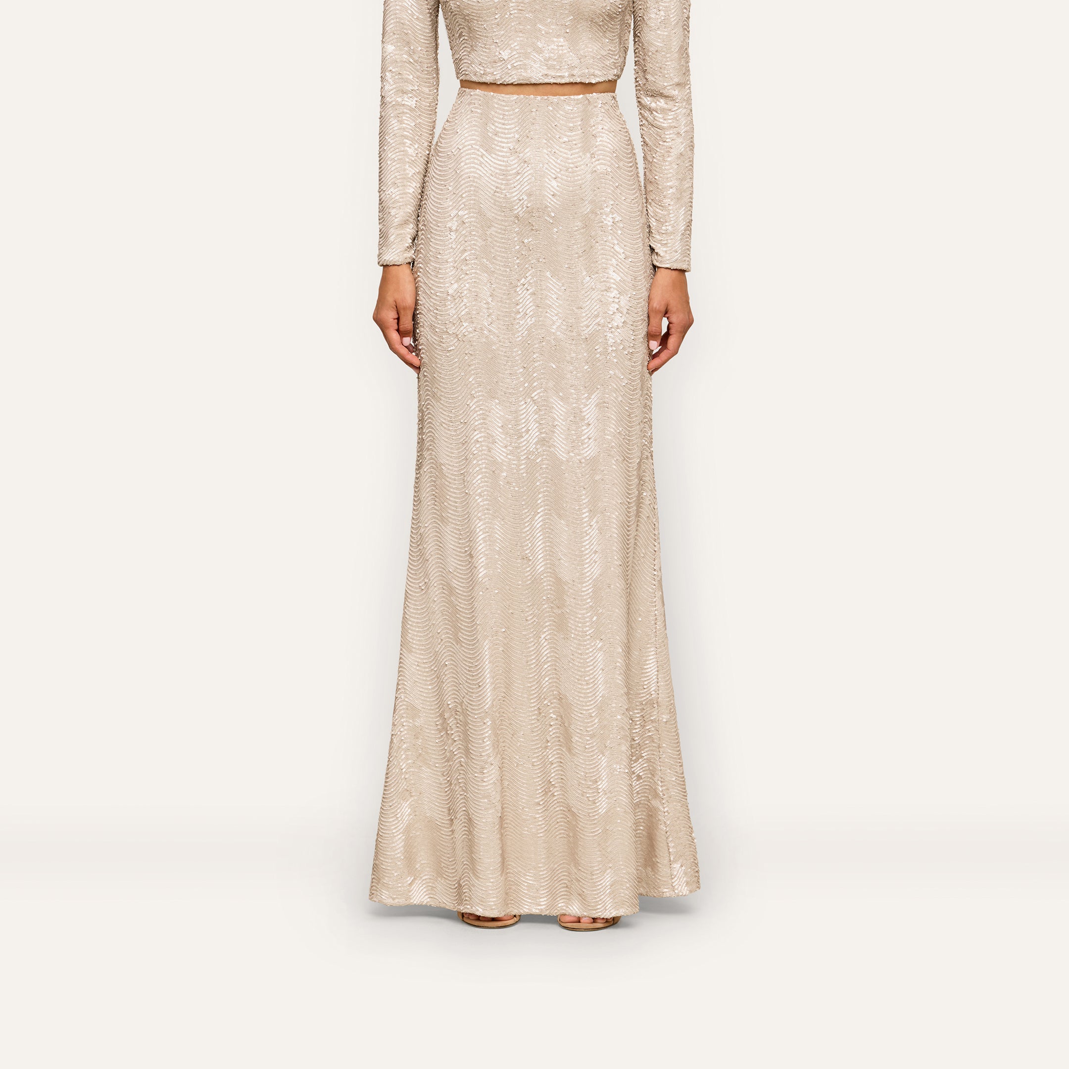 A person wearing the Zeta Sequin Maxi Skirt—a long, fitted, light beige skirt adorned with champagne sequins—stands against a plain white background. Only the body from the shoulders down is visible, making it a perfect look for a special occasion.