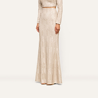 A woman stands against a plain background, her head out of frame, wearing the Zeta Sequin Maxi Skirt—a long, fitted skirt adorned with champagne sequins in a light beige hue—paired with a coordinating long-sleeved top for an elegant special occasion look.