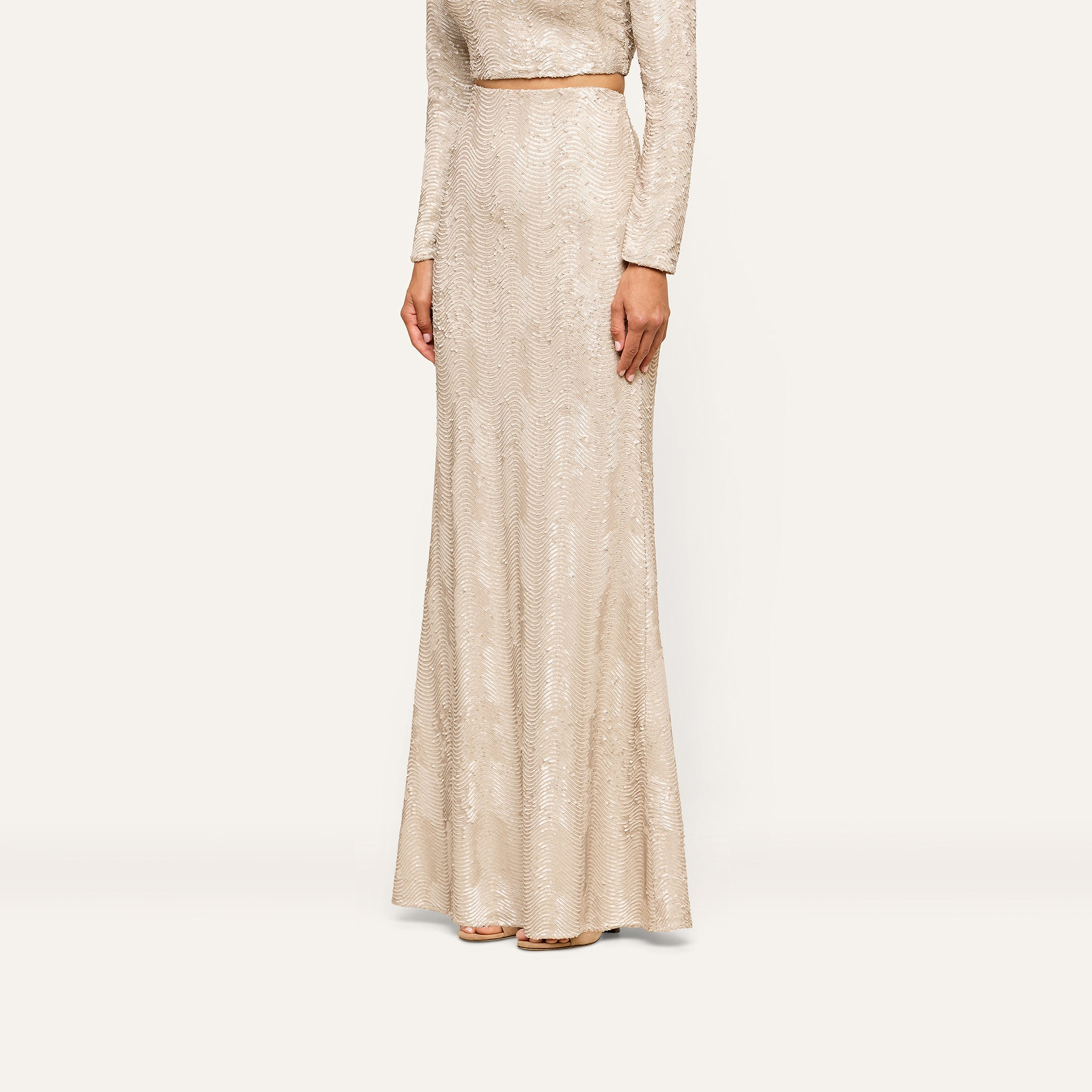 A woman stands against a plain background, her head out of frame, wearing the Zeta Sequin Maxi Skirt—a long, fitted skirt adorned with champagne sequins in a light beige hue—paired with a coordinating long-sleeved top for an elegant special occasion look.