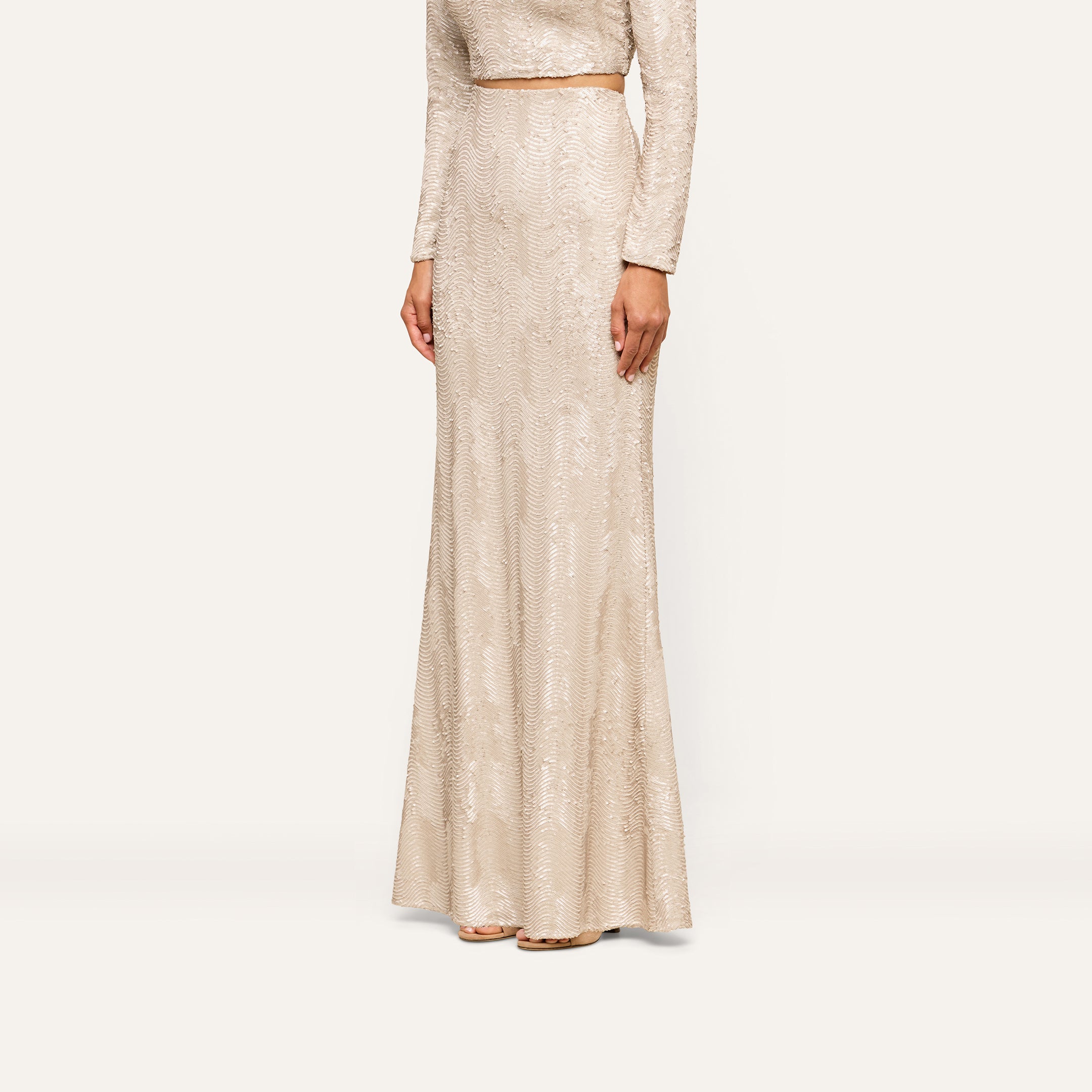 A woman stands against a plain background, her head out of frame, wearing the Zeta Sequin Maxi Skirt—a long, fitted skirt adorned with champagne sequins in a light beige hue—paired with a coordinating long-sleeved top for an elegant special occasion look.