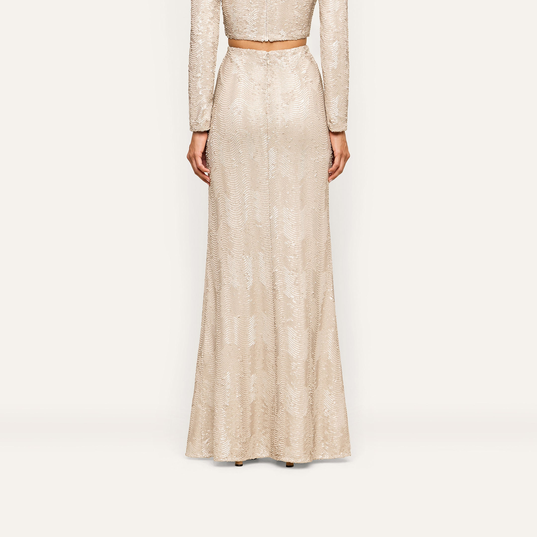 A person stands facing away, wearing the Zeta Sequin Maxi Skirt in champagne with a matching cropped top, both featuring a subtle shimmer—perfect for a special occasion.