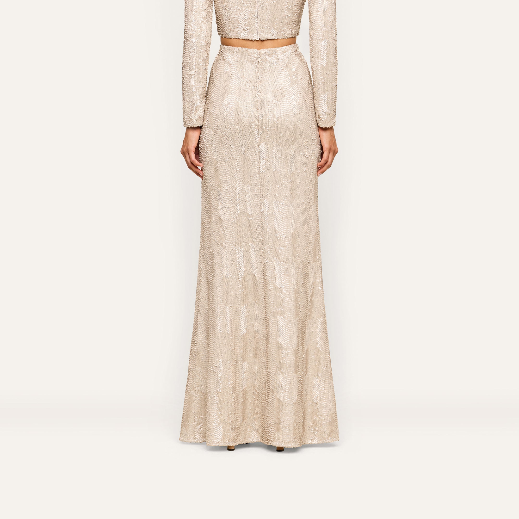 A person stands facing away, wearing the Zeta Sequin Maxi Skirt in champagne with a matching cropped top, both featuring a subtle shimmer—perfect for a special occasion.