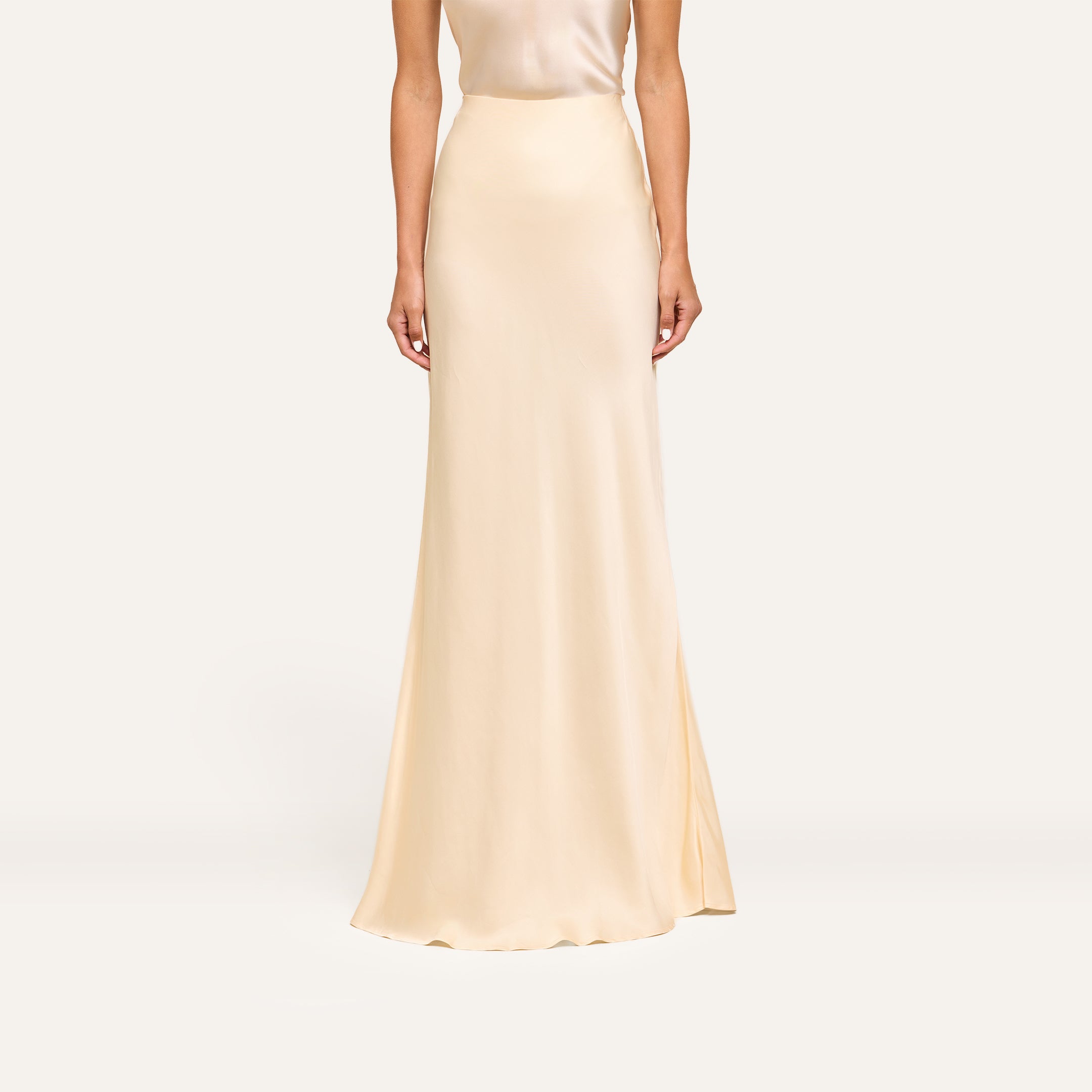 A woman wears the Zeta Satin Maxi Skirt, sleeveless and cream-colored, standing against a plain light background.