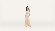 A woman stands against a plain white background, wearing a cream-colored satin tank top and a matching long skirt with beige heels. She has long brown hair and is accessorized with bracelets.